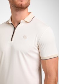Trumpas rankovėmis polo marškinėliai, pagaminti iš off-white spalvos medžiagos, su užtrauktuku. Dėlionėje yra tekstūruota apykaklė ir dryžių dekoracijos smėlio ir rudos spalvos. Pridėtas logotipo užrašas.