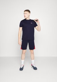 Conjunto deportivo de color azul marino con una camiseta de manga corta y pantalones cortos a juego que presentan acentos rojos a los lados. Se complementa con calcetines blancos y zapatillas azules.