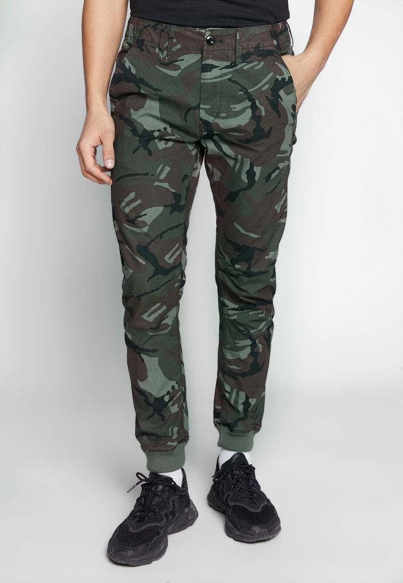 Kamouflage joggerbyxor i gröna och bruna nyanser, tillverkade av mjukt tyg, med elastisk midja och muddar vid anklarna, samt sidofickor.