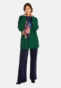 Lächelnde Frau in einem strukturierten grünen Cardigan, mit einem Blumenschal, blauen weit geschnittenen Hosen und schwarzen Schuhen, steht vor einem weißen Hintergrund.