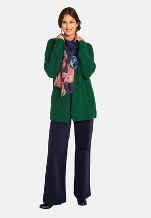 Lächelnde Frau in einem strukturierten grünen Cardigan, mit einem Blumenschal, blauen weit geschnittenen Hosen und schwarzen Schuhen, steht vor einem weißen Hintergrund.