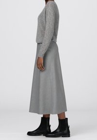 Henkilö, jolla on päällään harmaa neulepaita, harmaa midi-hame ja mustat nahkaiset nilkkasaappaat, seisoo sivuprofiilissa yksinkertaista taustaa vasten.