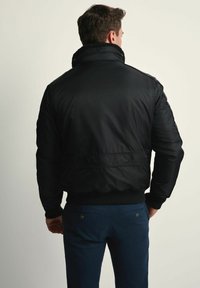 Mężczyzna ubrany w czarną kurtkę bomberkę z wysokim kołnierzem i niebieskie spodnie, stojący tyłem na jednolitym jasnym tle.