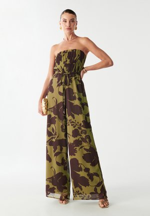 Kvinde iført stampefri olivengrøn jumpsuit med mørkt blomstermønster, som holder en guldfarvet clutch og bærer guldsmykker og hæle.