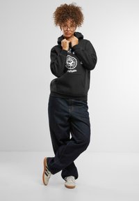 Juodas džemperis su baltu grafikiniu logotipu priekyje, derinamas su tamsiai mėlynomis džinsomis ir baltais sportbačiais, prieš šviesiai pilką foną.