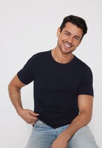 Uśmiechnięty mężczyzna o ciemnych włosach, ubrany w granatowy t-shirt i jasnobłękitne dżinsy, siedzący na tle jednolitej białej ściany.