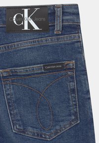 Džíny Calvin Klein s tmavě modrou denimovou texturou, viditelným šitím, nášivkou s logem CK a velkou zadní kapsou s jedinečným vyšívaným detailem.