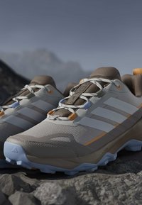 Paire de chaussures de randonnée beige et grises avec des accents orange sur un terrain rocheux, conçues pour une utilisation en sentier extérieur.