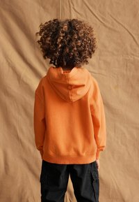 Felpa arancione con tasca a marsupio, polsini e orlo a coste, abbinata a pantaloni cargo neri. Tessuto di cotone morbido, vestibilità ampia, cappuccio decorativo.