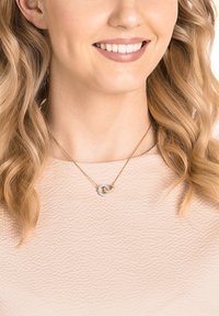 Roosa kuldne ripatsikaelakee, millel on omavahel põimunud silmused, sisaldab väikseid selgeid gemme. Peen ahel tekstuuriga heledal ülemisel.