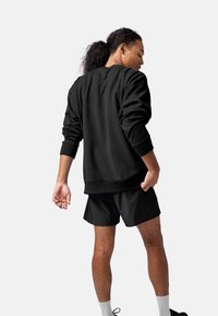 Felpa nera oversize con polsini a coste, abbinata a pantaloni corti neri. Tessuto morbido; modello visto da dietro, in piedi in modo casual.