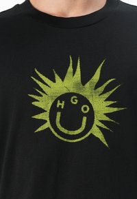 Black cotton t-shirt with round neckline featuring a large yellow-green graphic resembling a sun with a smiley face and the letters "HGO."