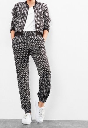 Personne portant une veste zippée à motifs noirs et blancs assortie à un pantalon et des baskets blanches, debout contre un mur blanc.