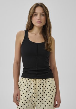 Jeune femme aux longs cheveux bruns portant un haut noir sans manches et un pantalon crème à pois noirs sur un fond gris.