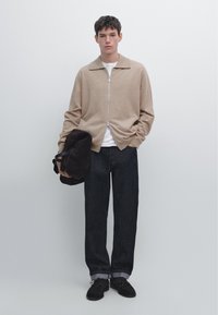 Ung man som bär en beige zip-up tröja, vit skjorta, mörka jeans, svarta skor, håller en svart väska mot en enfärgad ljus bakgrund.
