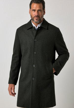Homme portant un long manteau gris foncé boutonné, une main dans la poche, debout devant un fond clair uni.