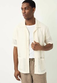 Camisa de botón de manga corta en color crema con un diseño de punto abierto texturizado. Presenta un cuello relajado y dos bolsillos frontales. Combinada con pantalones beige.