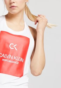Valkoinen hihaton paita, jossa on suuri punainen logo "CALVIN KLEIN PERFORMANCE". Sileä puuvillakangas, urheilullinen taakse avautuva malli, minimalistinen tyyli.