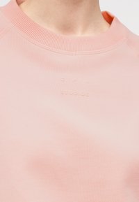 Κοντινό πλάνο ενός ανοιχτό ροζ φούτερ με στρογγυλή λαιμόκοψη και διακριτικό ανάγλυφο κείμενο στην περιοχή του στήθους που λέει «CPH STUDIOS».