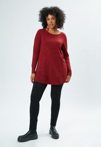 Bordeauxrote langärmelige Strick-Tunika mit einem dezent strukturierten Muster, kombiniert mit schwarzen Leggings und klobigen schwarzen Stiefeletten.