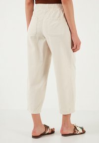 Light beige cropped pants with an elastic waistband, two back pockets, and a textured fabric. Worn with brown flat sandals featuring embellishments.