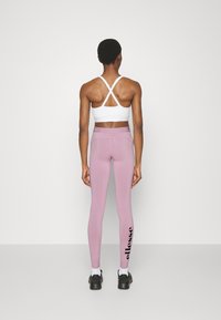Idrottslig person som bär en vit sport-bh och rosa leggings med "ellesse"-logo, stående vänd mot en enkel ljusgrå vägg.