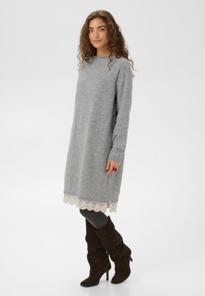 Une femme porte une robe-pull grise en maille avec un ourlet en dentelle blanche, des collants gris et des bottes marron foncé jusqu’aux genoux, sur un fond uni.