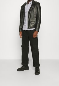 Giacca da motociclista in pelle nera con accessori in argento, camicia grigia con bottoni, pantaloni cargo neri con tasche laterali e stivali neri con lacci.