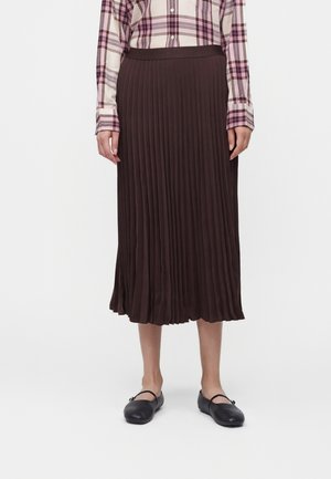 Personne portant une jupe midi plissée marron, des chaussures Mary Jane noires et une chemise à carreaux violette et blanche à manches longues avec boutons.