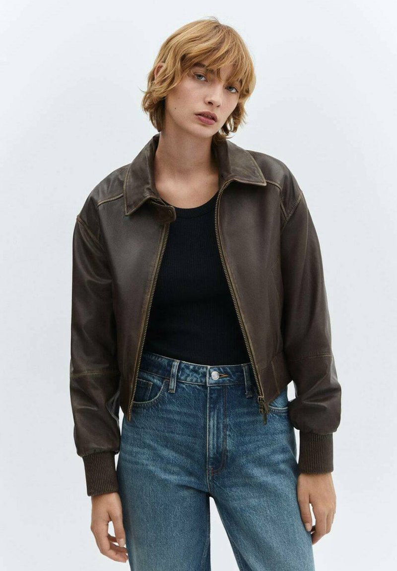 Braune Lederbomberjacke mit Kragen, Reißverschluss und gerippten Bündchen, getragen über einem schwarzen Oberteil und kombiniert mit Jeans.