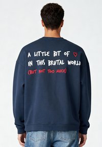 Tumši zila sporta krekls ar apaļu kakla izgriezumu, aizmugurē ir balti un sarkani uzraksti. Mīksta, teksturēta auduma, ar ribotām aprocēm un apakšmalu.