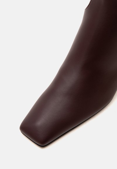 Gros plan d'une botte en cuir brun foncé avec une surface lisse et un bout carré sur un fond clair.