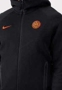 Sudadera con cremallera negra, fabricada en tejido suave, que presenta un logotipo de Nike en color naranja y un emblema redondo naranja. Incluye bolsillos laterales con cremallera.