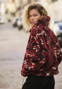 Kurtka polarowa w kolorze bordo i kremowym z kwiatowymi wzorami, wysokim kołnierzem, elastycznymi mankietami i luźnym krojem, noszona na czarnych spodniach.