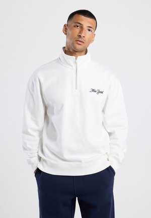 Sudadera blanca con cremallera y cuello alto, que presenta un pequeño detalle bordado en negro con la palabra "New York" en el lado izquierdo del pecho. Textura suave.