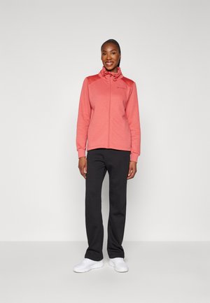 Chaqueta rosa claro con cremallera y cuello alto, pantalones negros y zapatillas blancas. La chaqueta presenta una textura suave y un detalle del logo en el pecho.