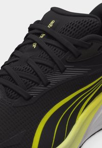 Μαύρο αθλητικό παπούτσι με μαύρα κορδόνια, κίτρινο και μαύρο κυρτό σχέδιο στο πλάι και αναπνεύσιμο επάνω μέρος από δίχτυ, σε κοντινή λήψη.