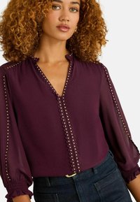 Vīna sarkanā blūze ar viļņotu kakla izgriezumu, garām caurspīdīgām piedurtēm un zelta kniedēm gar priekšējo daļu. Gluda auduma, pieguļoša vidukļa daļā.