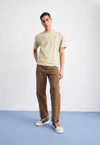 Camiseta de algodón de color beige claro con una textura sutil, combinada con jeans rectos marrones y zapatillas verde olivo con detalles en crema.