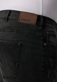 Jeans de mezclilla negra con una etiqueta de cuero marrón que dice "MEN+". Presentan un diseño elegante, bolsillo lateral y un mínimo desgaste en la tela.