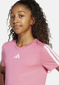 Camiseta deportiva de manga corta en color rosa, con cuello redondo y tres rayas blancas en cada hombro. Presenta un logo blanco en el pecho.
