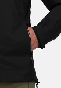 Svart utomhusjacka med en texturerad yta, utrustad med en sidoficka med dragkedja och justerbara ärmslut. Materialet verkar lätt och hållbart.