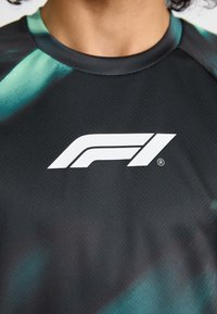 Zwart sportshirt met een verlooppatroon in teal. Bevat een wit F1-logo op de borst. Gemaakt van textuurstof die ademend is.