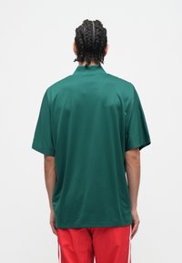 Asmuo su pintais plaukais, vilkintis žalią trumpomis rankovėmis marškinėlį ir raudonas kelnes, atsukęs nugarą priešai paprastą baltą foną.