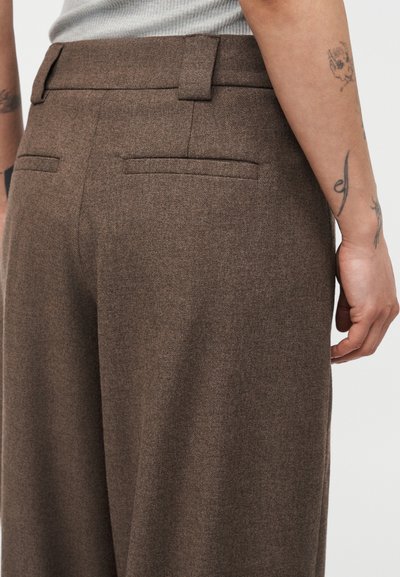 Καφέ μάλλινο παντελόνι με πίσω τσέπες welt, που φοριέται από ένα άτομο με τατουάζ στα χέρια και ελαφρύ γκρι τοπ μέσα στην μέση.