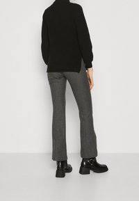 Osoba ubrana w szare spodnie w jodełkę, czarny sweter i czarne botki, stojąca tyłem na tle białej ściany.