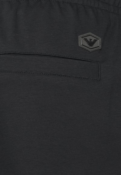 Shorts noirs en tissu texturé, avec une taille élastique et un badge logo hexagonal cousu sur la poche latérale gauche.
