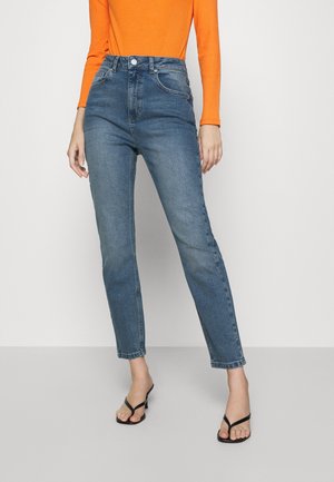 Frau in hochtaillierten blauen Jeans, langärmligem orangen Rippoberteil und schwarzen Absatzsandalen, die vor einem schlichten Hintergrund steht.