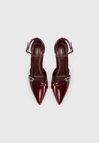 Μπορντό μυτερές γόβες από δερματίνη με λουράκι στον αστράγαλο, που διαθέτουν διακοσμητικό αγκράφα στο λουράκι και γυαλιστερό φινίρισμα.