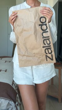 Fehér ruhát viselő személy egy gyűrött barna Zalando bevásárlótáskát tart a törzse előtt, zárt térben.